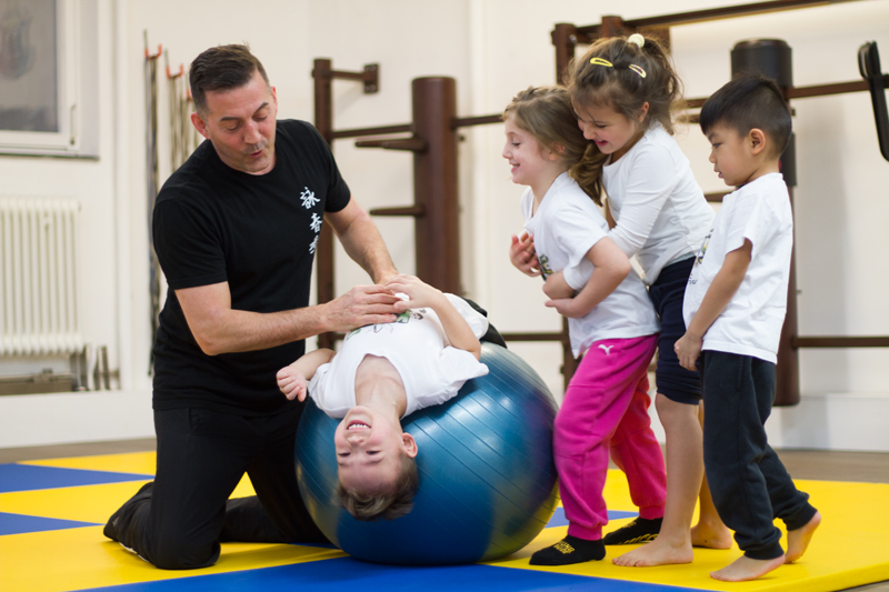 Vier Kindern: Eines lehnt sich rückwärts über einen großen Gymnastikball. Trainer gibt Hilfestellung