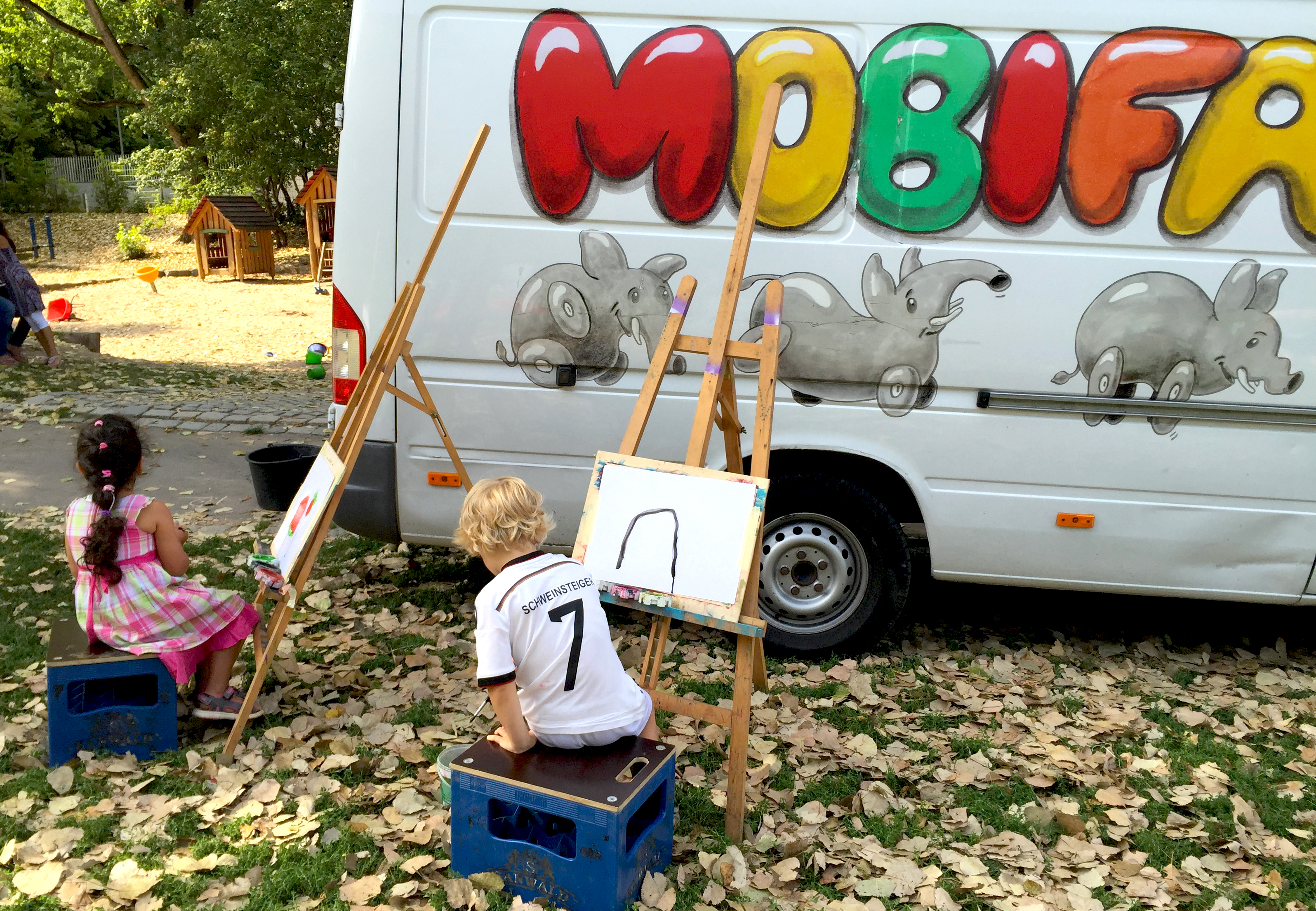 © stjg Zwei Kinder sitzen je vor einer Staffelei, dahinter der Mobifant Bus.