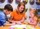 Eine junge, blonde Frau sitzt mit zwei kleinen Kindern an einem bunten Tisch in der Spiele-Erfinder-Werkstatt der Ravensburger Kinderwelt Kornwestheim und malt mit ihnen.
