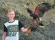 In der Falknerei auf der Burg Guttenberg steht ein blonder Junge mit schwarz-weißem T-Shirt vor einer Felswand. Er trägt einen dicken, großen Lederhandschuh und ein braun-schwarzer Greifvogel mit orangefarbenem Schnabel landet auf seinem behandschuhten Arm