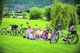 Mehrere gutgelaunte Eltern und Kinder sitzen in Heidelberg umringt von Fahrrädern mit einem geflochtenen Korb auf einer karierten Picknick-Decke.