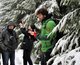 Ein Mann in grüner Winterjacke steht zwischen schneebehangenen Bäumen. Im Hintergrund sind weitere Menschen vor verschneiten Tannenbäumen zu sehen.
