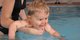 Kleinkind und Mutter befinden sich in einem Schwimmbecken. Die Mutter hält das Baby über Wasser.