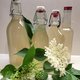 Vier Flaschen mit Holundersirup, davor ein paar Holunderblüten.